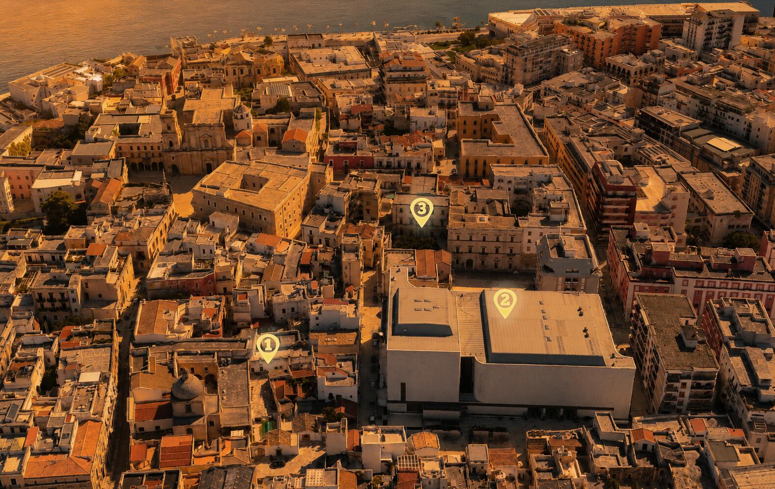 Veduta aerea al tramonto del centro storico di Brindisi affacciato sul porto: tre segnaposto luminosi indicano i poli del parco archeologico diffuso nell'area tra Via Duomo e San Pietro degli Schiavoni.