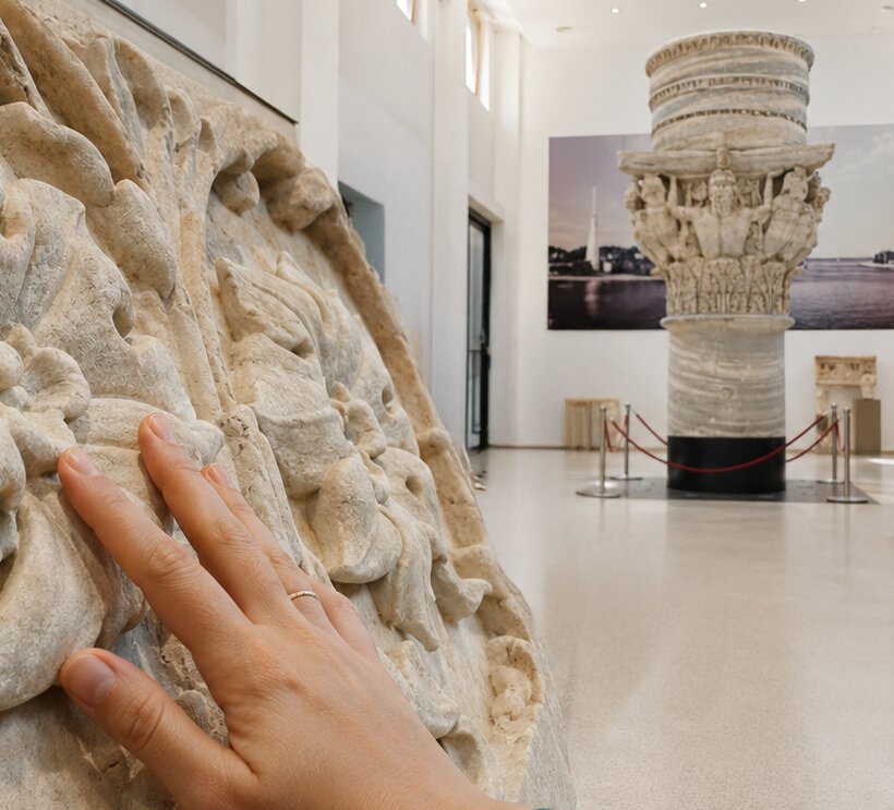 Una mano esplora con le dita un rilievo marmoreo di età romana in primo piano; sullo sfondo, la sala museale con il capitello della colonna romana di Brindisi e statue esposte su basamenti.